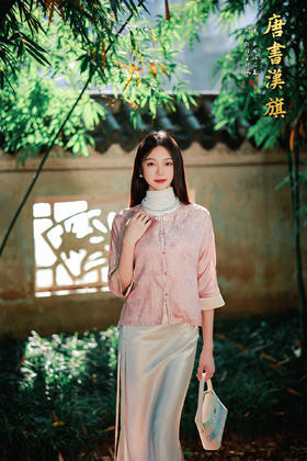 【丝语花痕(人丝提花）】粉调提花短款衬衣，融国风雅致于现代剪裁。   衣身采柔粉提花面料，暗蕴细腻纹理，圆领设计衬得颈部线条温婉；精致花扣点缀门襟，七分袖口饰浅香槟色滚边，细节处尽显复古巧思。